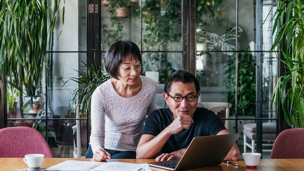 A middle aged man using laptop with a woman looking over shoulder and documents spread on the table.