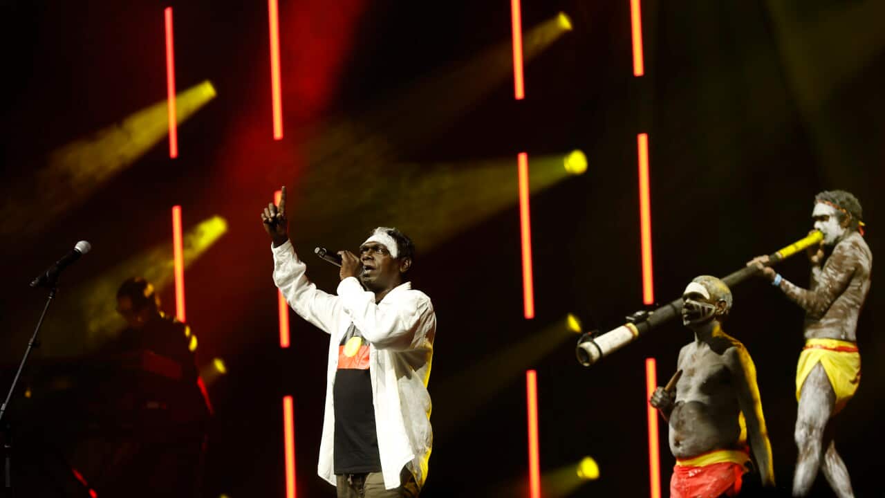 An Aboriginal Australian singer in a white long-sleeved collared shirt points his finger upwards on a dark stage with red and yellow lights, while three men in body paint and red and yellow loincloths perform beside him, one playing a didgeridoo.