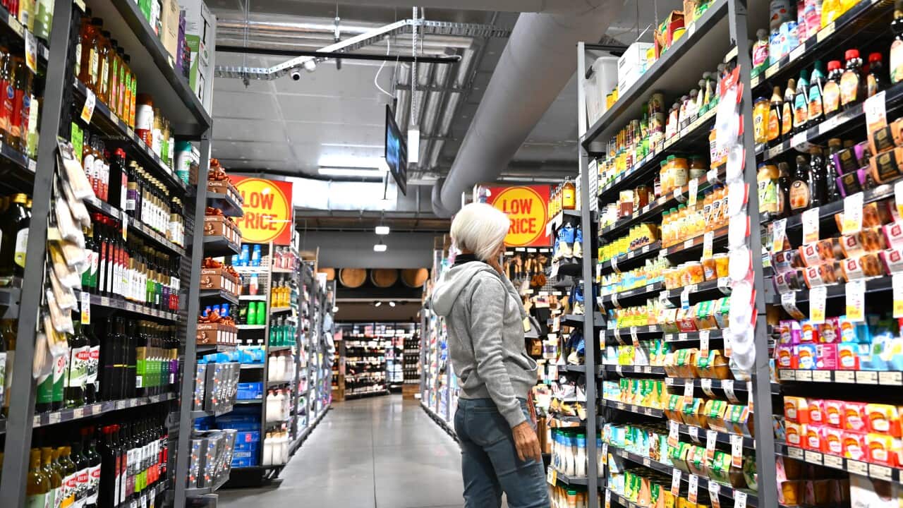 A woman with grey hair wearing a grey sweater and jeans looks at items in an aisle of a supermarket.