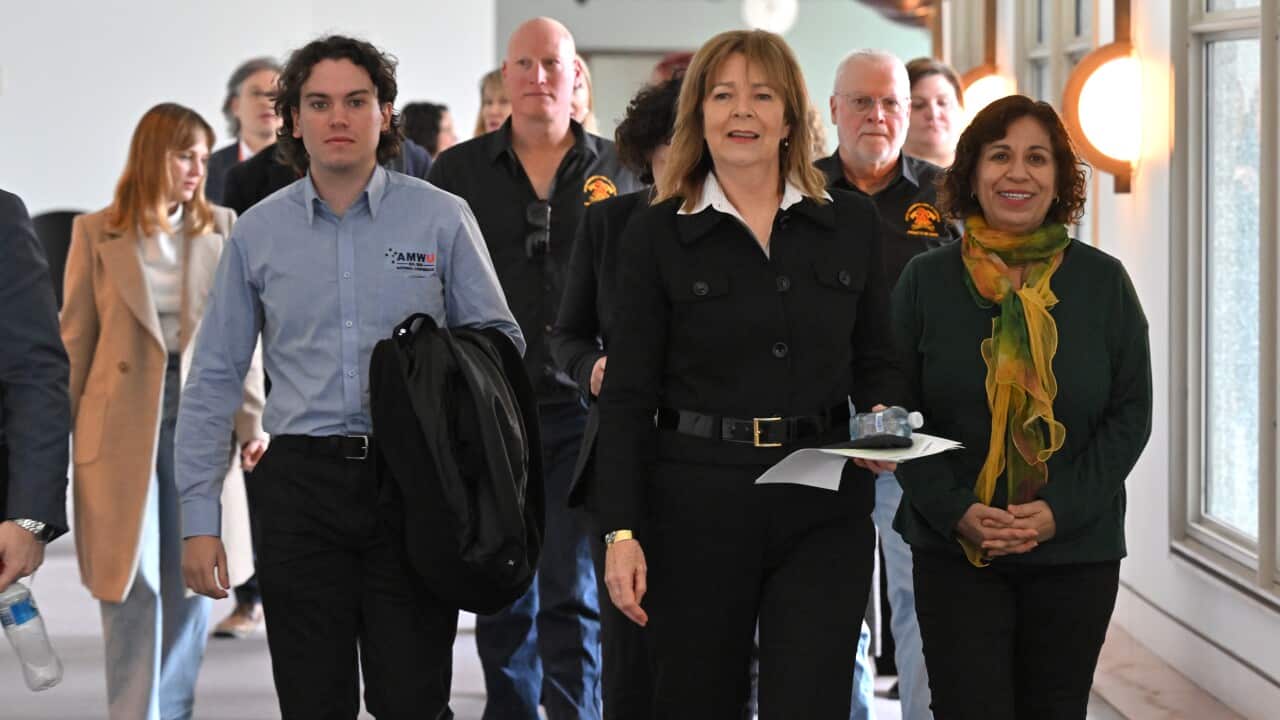 A group of people walk down a hallway, in the lead is a woman in a black shirt and pants carrying a piece of paper