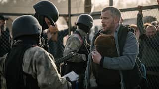 A man stands in front of a chain fence, talking to three people in soldier's uniforms. 