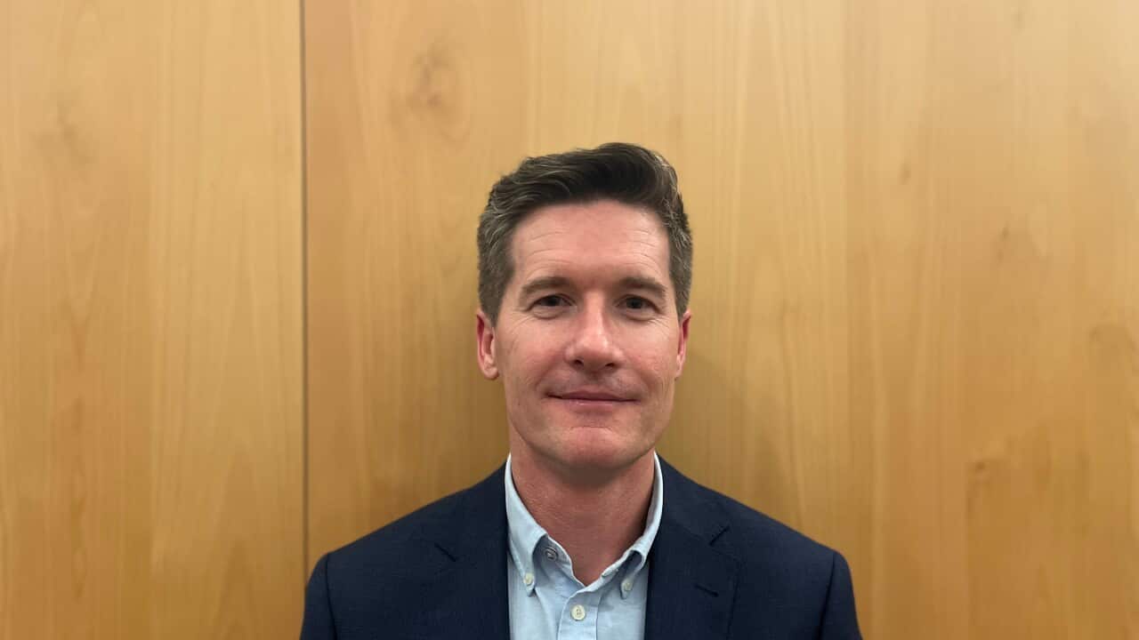 a middle aged man in a suit looks neutrally at camera in front of a plain wooden background
