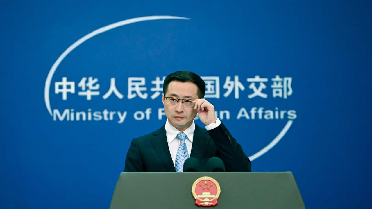 A man in a suit and glasses stands at a podium featuring the national emblem of China, speaking in front of a blue backdrop for the Ministry of Foreign Affairs.