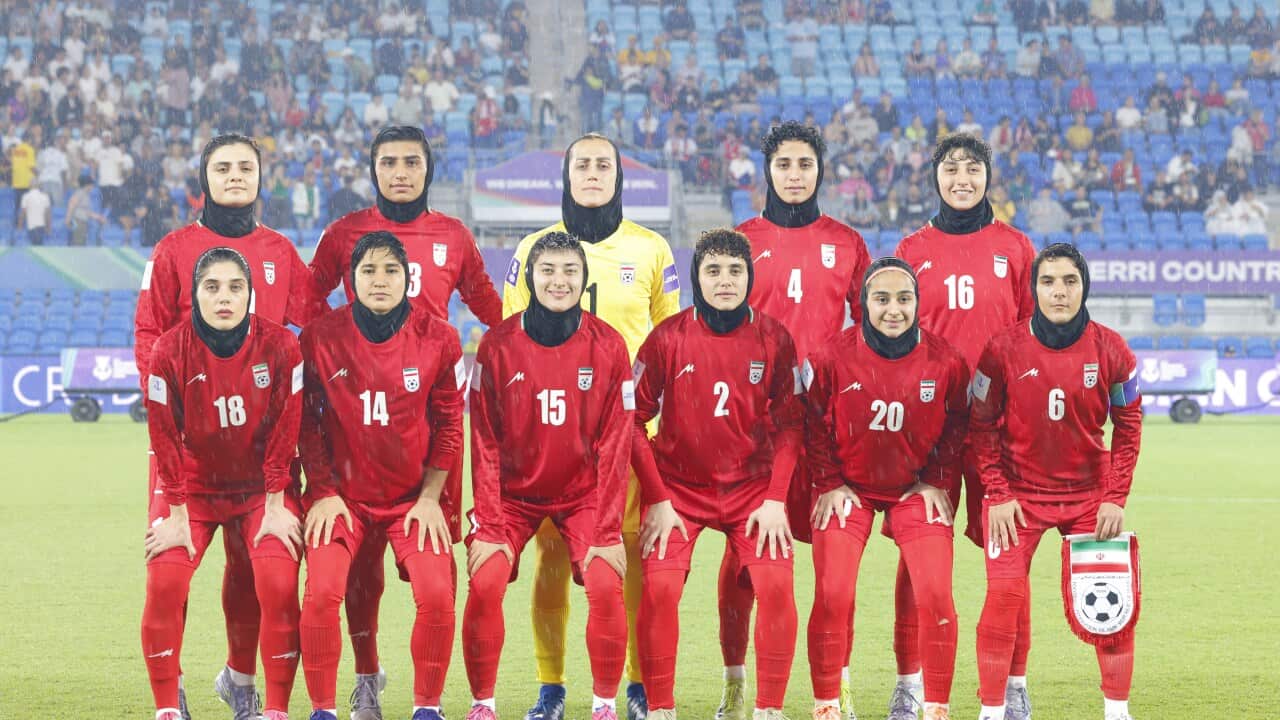 A team of female soccer players from the Iranian national team, wearing red kits and black hijabs, poses for a group photo on a rainy pitch before a match.