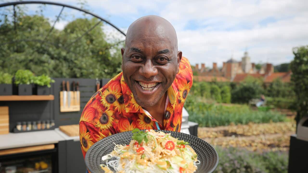 Wearing a bright shirt, Ainsley Harriott stands in an outdoor kitchen leaning forward to present a plate of food.