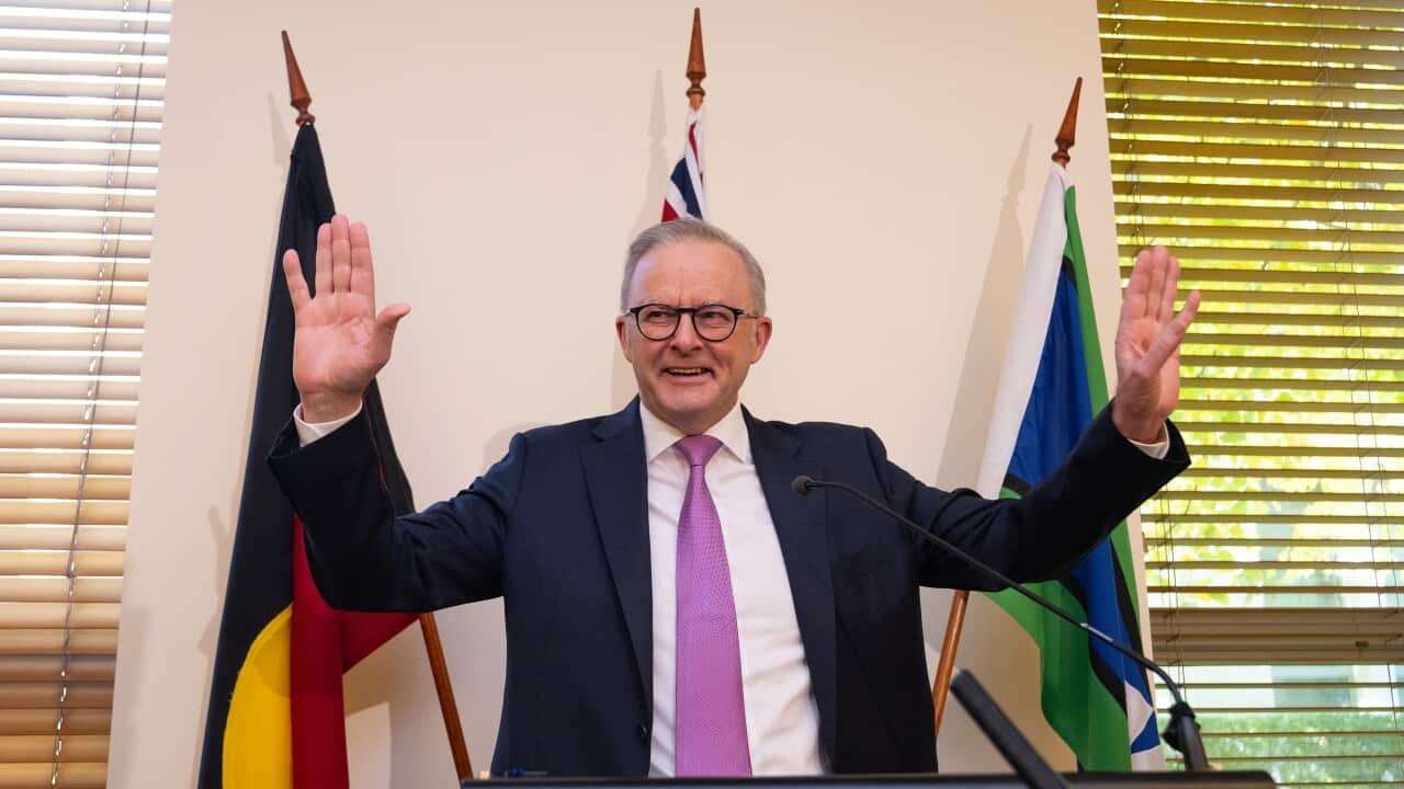 A man in a dark suit, white shirt and purple tie smiles and holds his hands up in front of three flags
