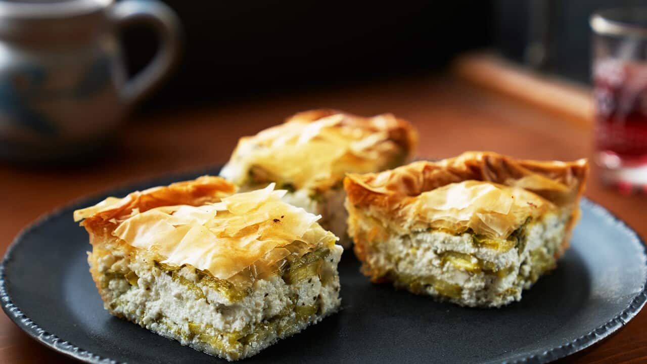 Three pieces of pie, with a cream filling studded with green and flaky golden filo pastry o top, sit on a dark round plate on a table. A mug and glass can be see, out of focus, in the background.