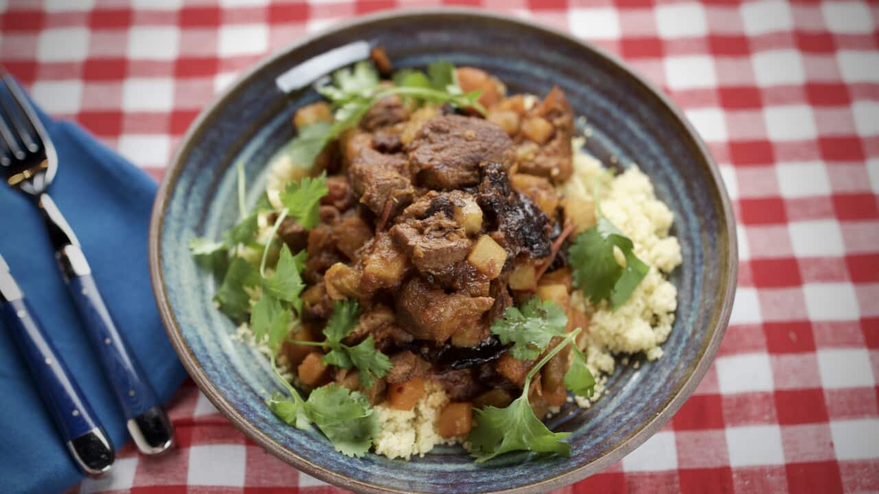 A rich meat tagine sits on a bed of couscous in a blue-grey pottery bowl, on a red-checked table cloth.