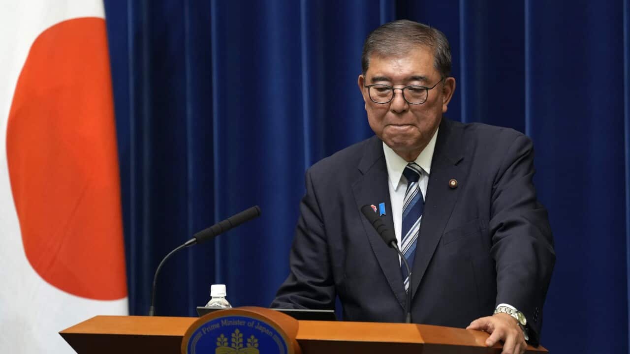 A man in a blue suit and striped tie stands behind a podium, his face expressing sadness.