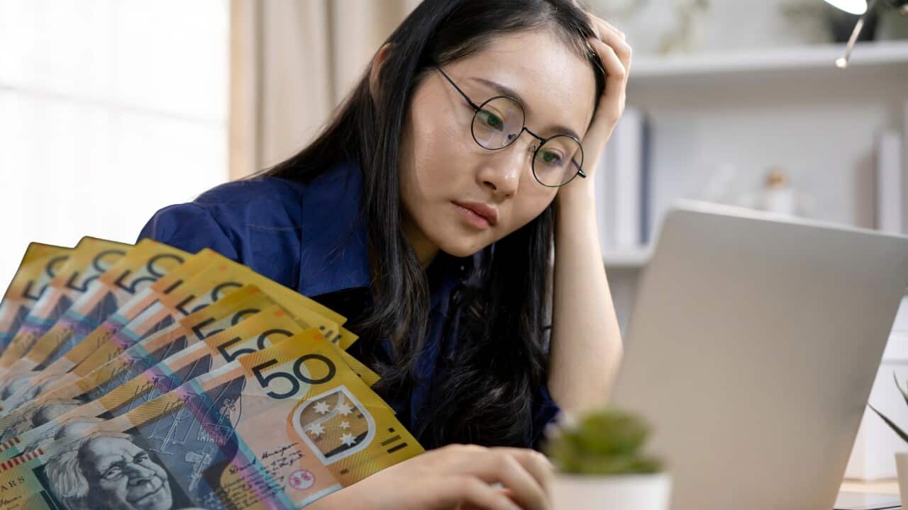 A frustrated young woman with glasses sits at a desk, looking at a laptop with a pained expression She rests her head in one hand, appearing stressed or overwhelmed by her finances. Superimposed to her right is a fan of fifty-dollar Australian banknotes. 