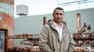 A man in a pink collared shirt and fawn-coloured slouchy jacket stands in front of graffitied rusting pipes, with a wall behind.