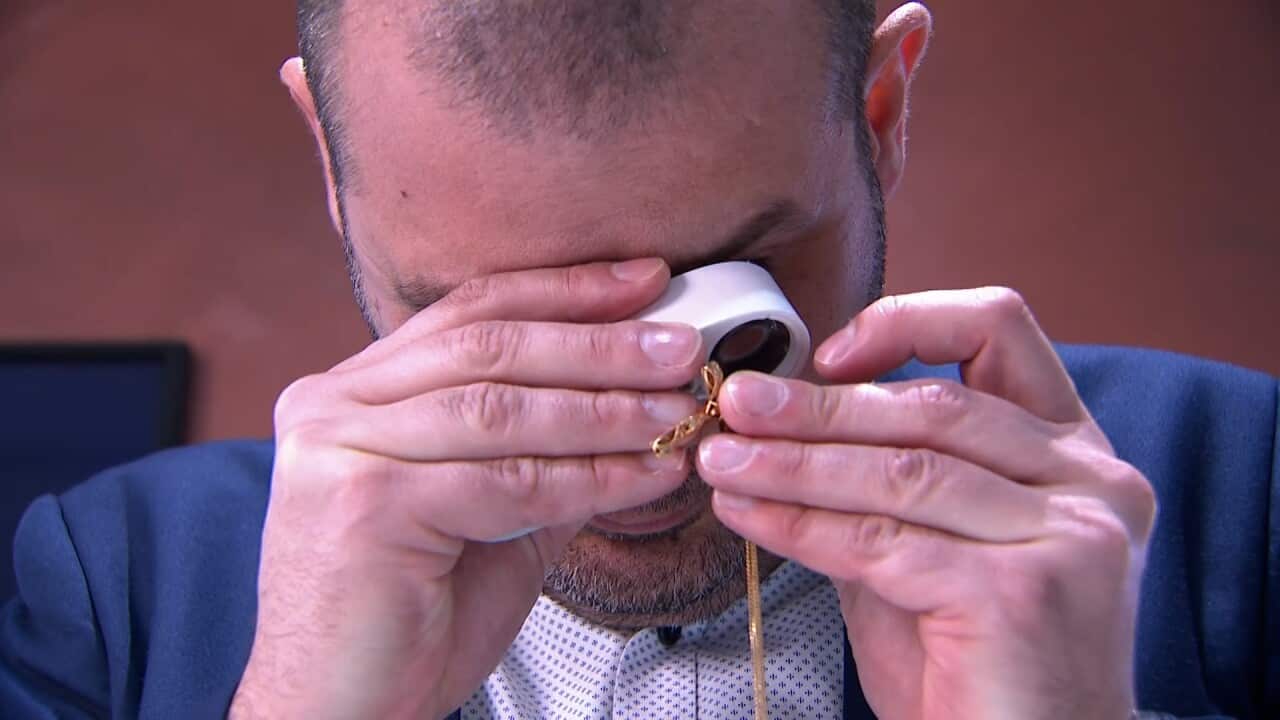 A man in a suit jacket and a collared shirt uses a jeweller's loupe to inspect a piece of gold jewellry.