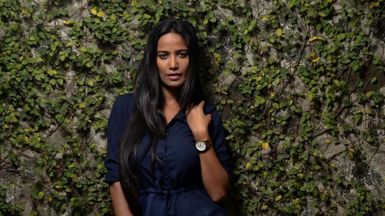 Poonam Pandey standing against a wall of green foliage.
