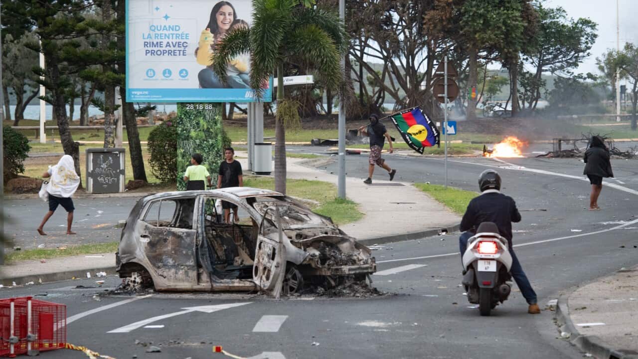 A burnt out car and protesters and rioters walking on the streets.