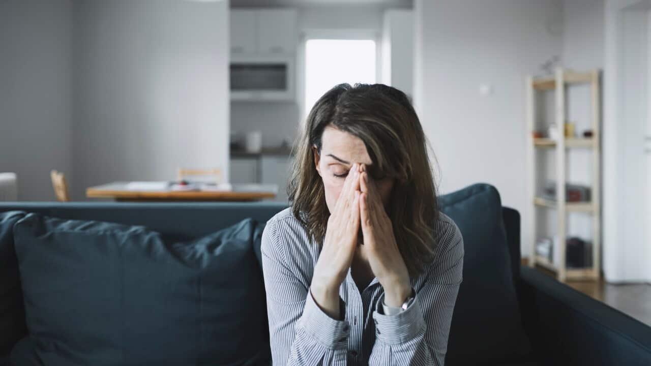 A woman is sitting on a couch with her hands on her face. She has a look of distress on her face.