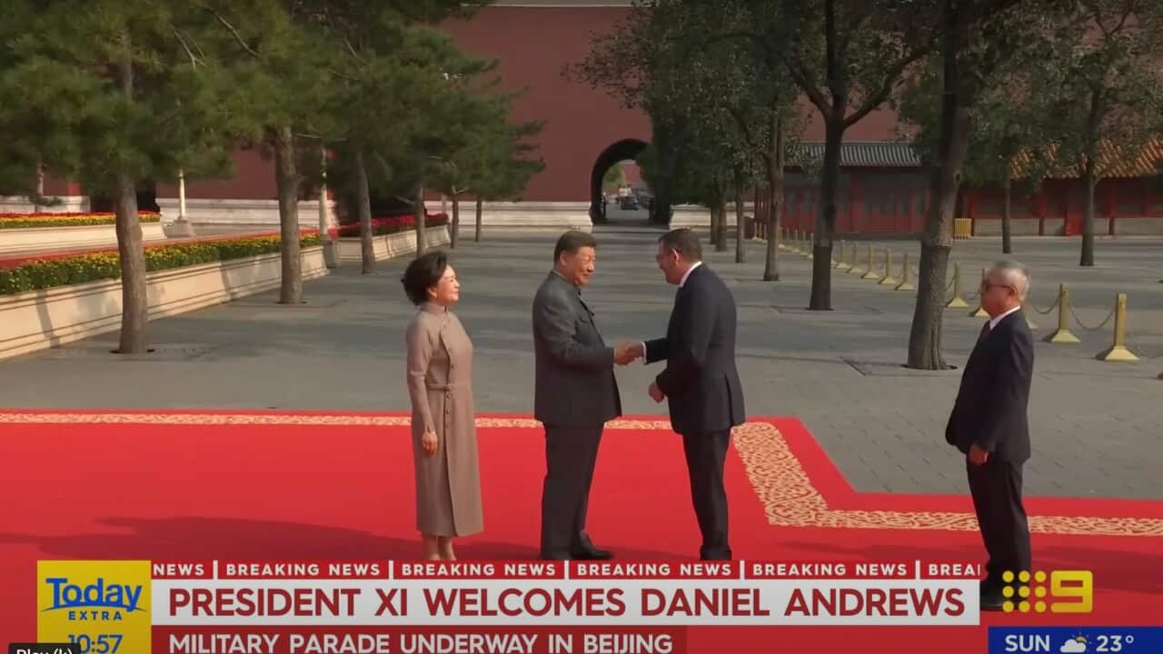 Two men in suits shake hands as they stand on a red carpet. There's a woman and a man standing at some distance.