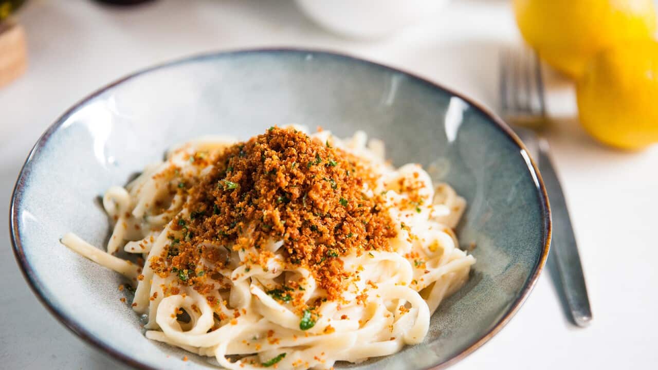 A mound of pasta with a light creamy sauce sits in a wide pottery bowl, topped with dark golden breadcrumbs.