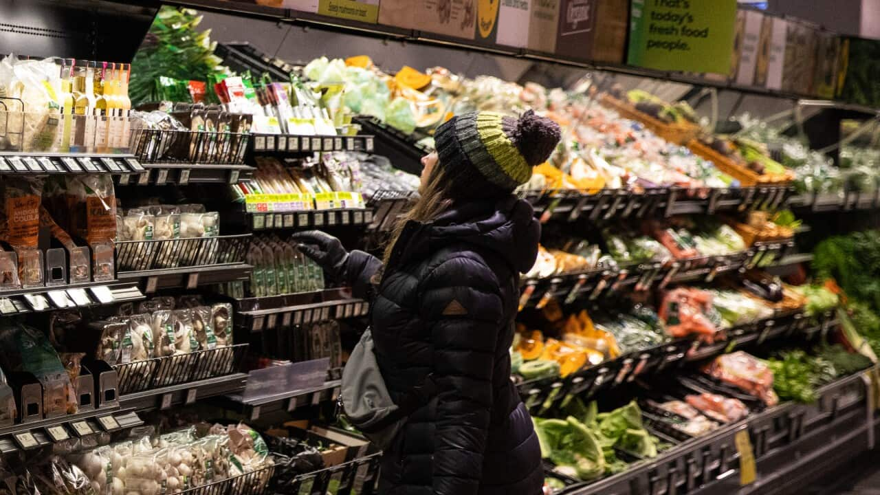 A woman in a beanie and black coat plucks an item from a vegetable aisle in a supermarket.