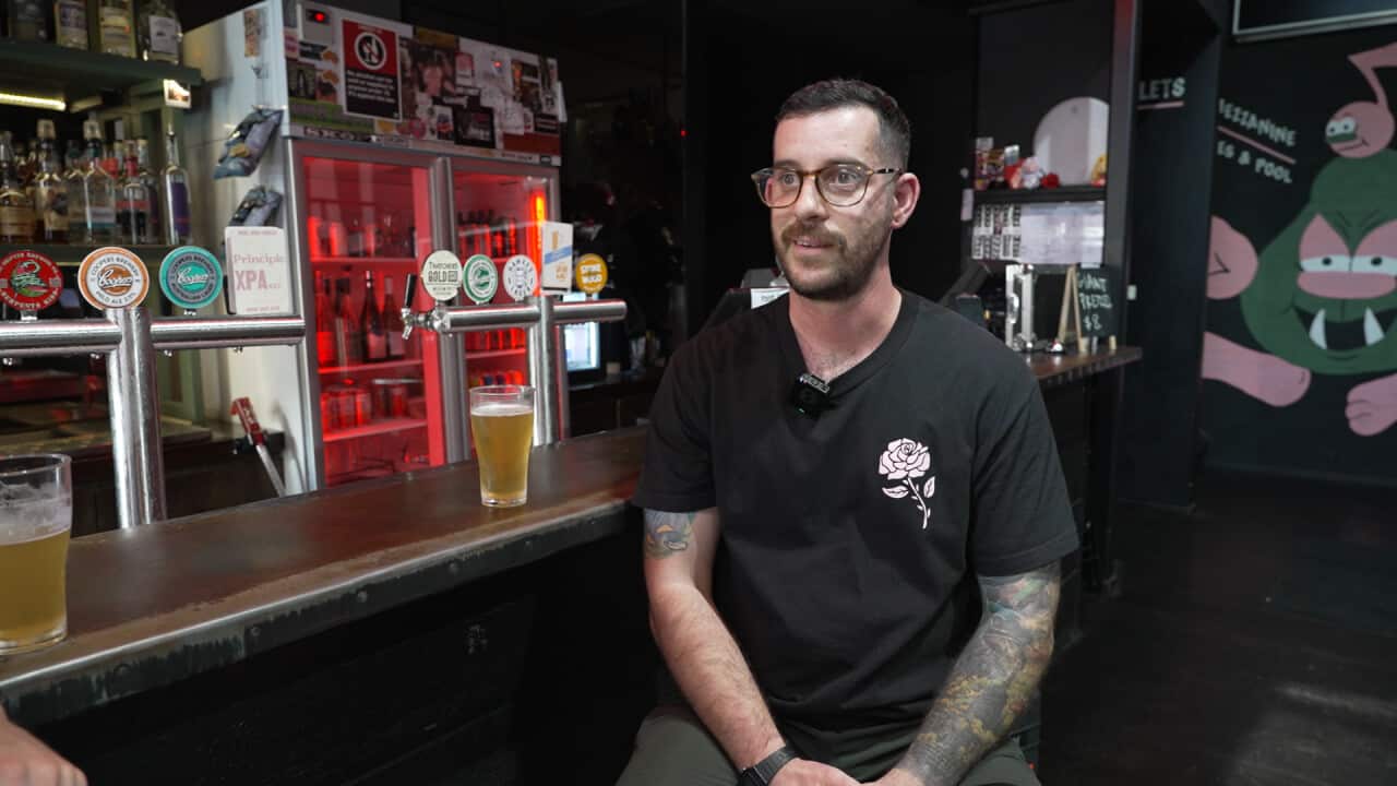 A man with a beard and arm tattoos is sitting at a bar with two beers on the bench next to him