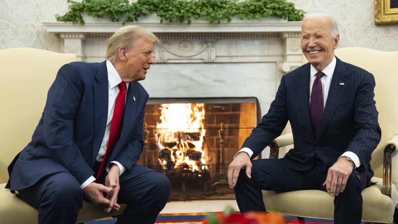 Two men in suits sitting and smiling in front of a lit fireplace.