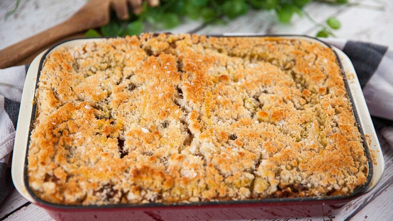 A deep red cast iron baking dish sitting on a white wooden surface is seen from overhead. It has a baked filling, topped with golden breadcrumbs.