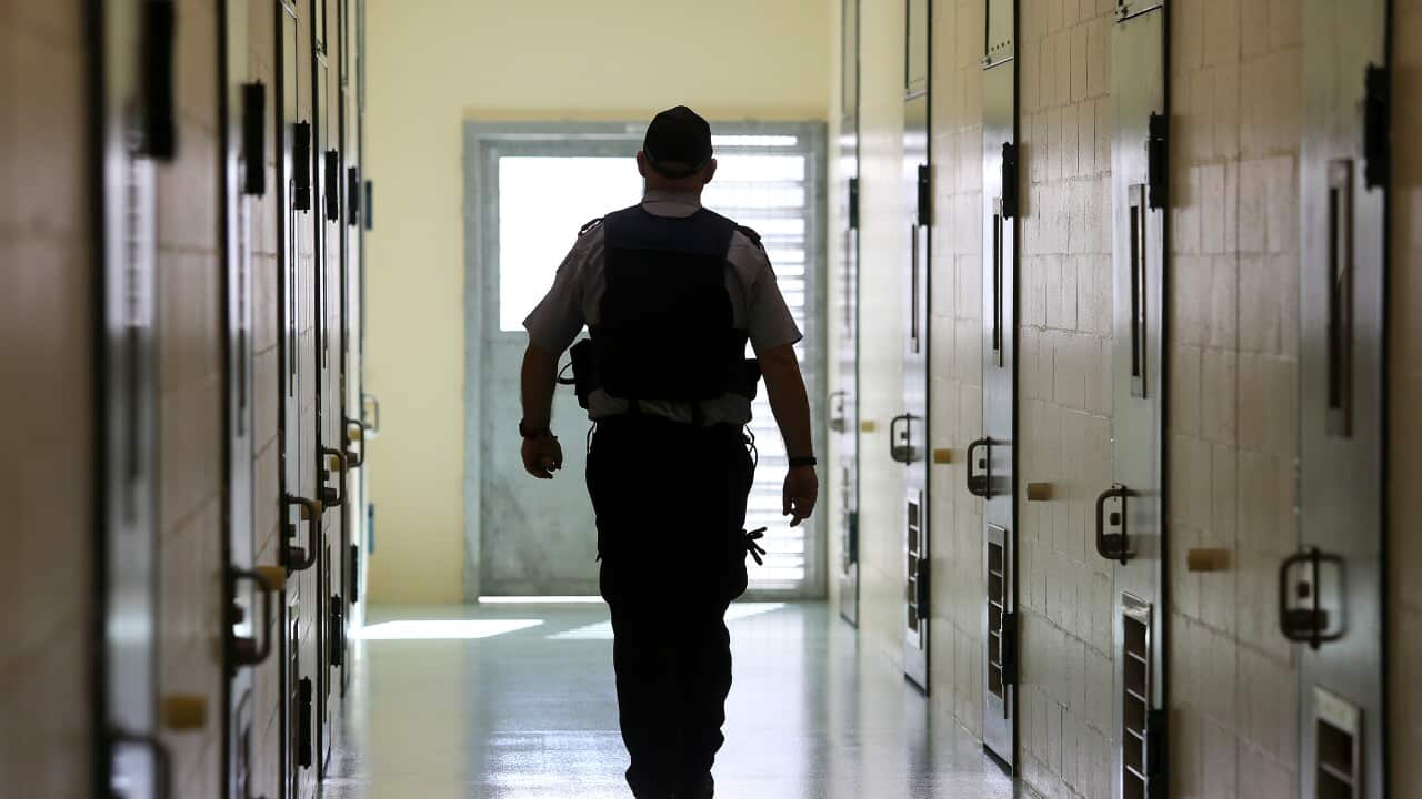 A prison guard walking down a corridor with cells either side of him.