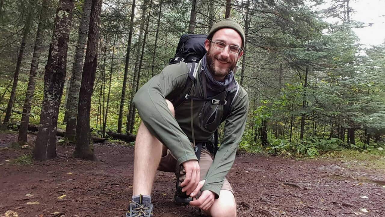 A white man wearing outdoor gear and a backpack kneels in some woods.