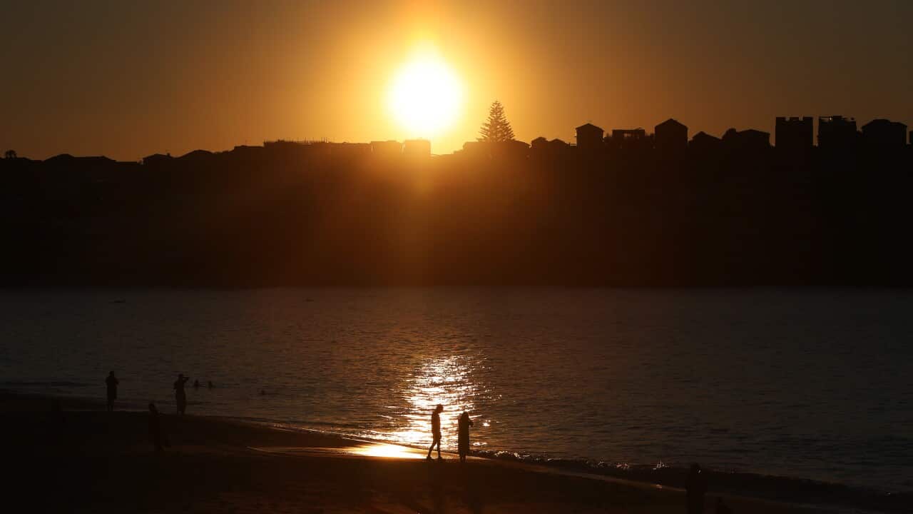 A dark image with a bright orange sun setting over a beach landscape, there are two people in the foreground but the focus is on the bold red sun.