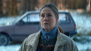 A worried woman is seen, from the waist up, standing in front of a car in a snowy landscape.