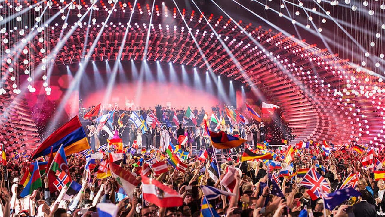 Crowds of spectators stand in front of a brightly lit Eurovision stage, waving national flags from around the world.