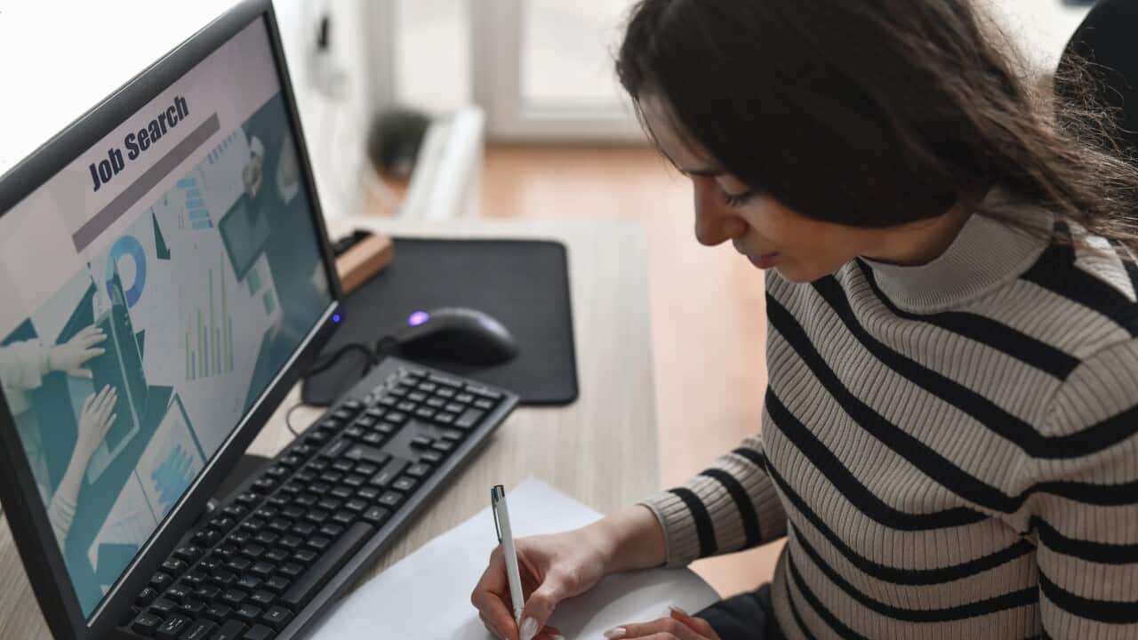 A woman sitting at a desk, writing on a piece of paper. A job search website is open on the computer screen in front of her