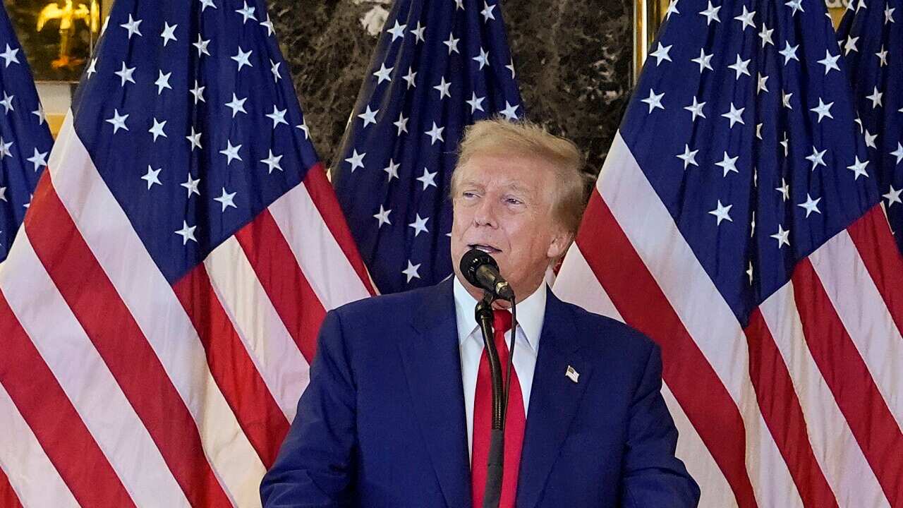 Donald Trump speaking into a microphone in front of a row of American flags.