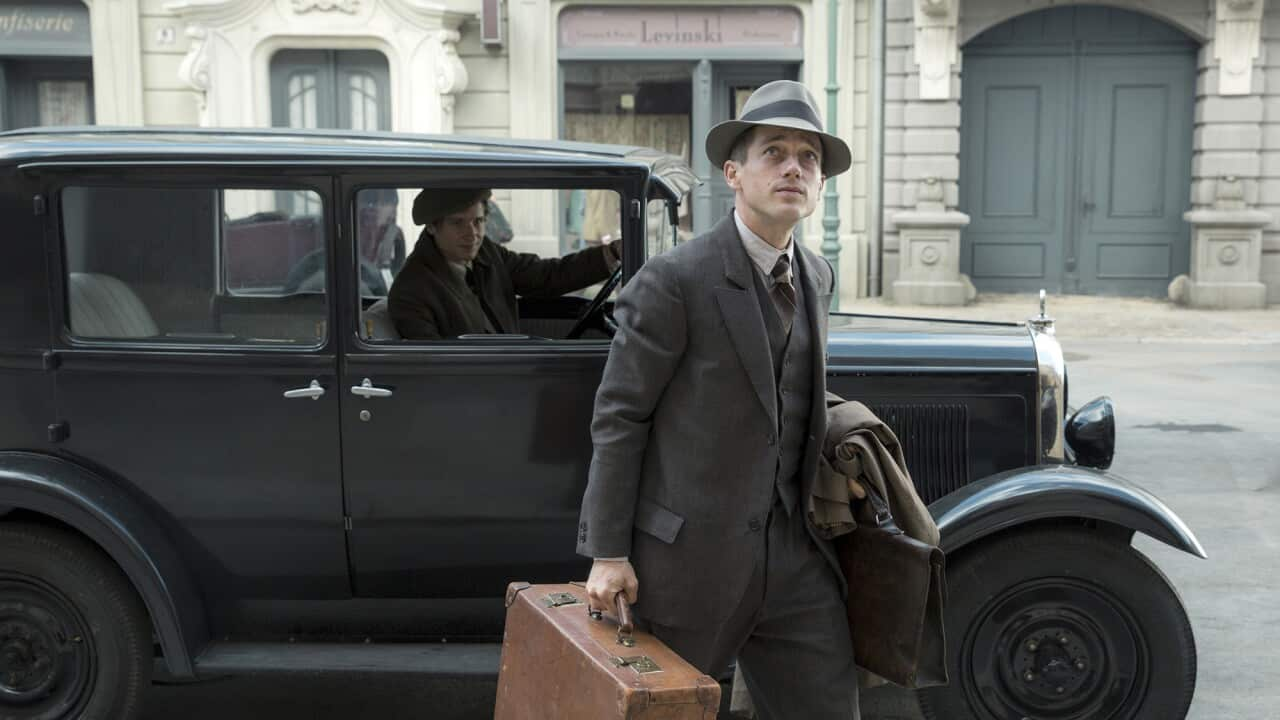 A young man in a 19020s-style suit and hat, carrying a suitcase, stands in front of a car, looking upwards. 