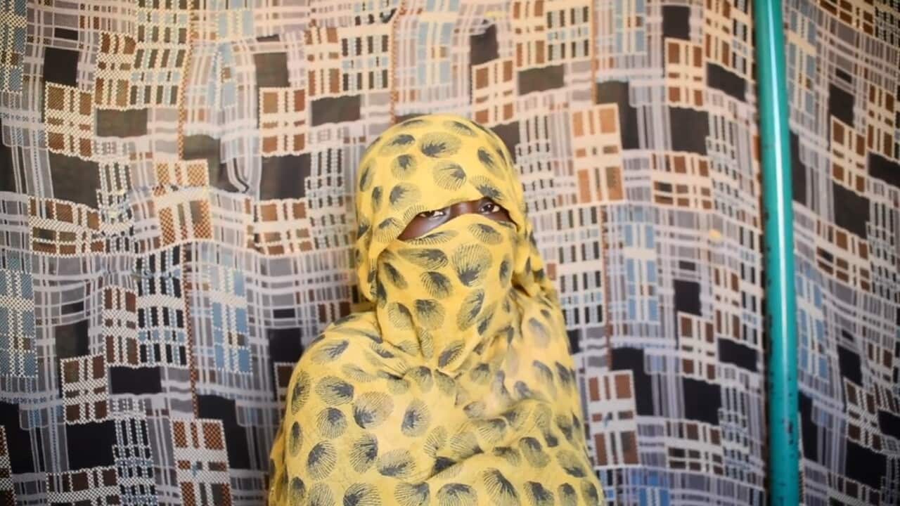 A woman sitting in front of a patterned fabric backdrop that features a repeating geometric design in shades of tan, brown, white, and black. She is wearing a bright yellow face covering with a black spotted or feathered pattern, which covers her entire head and face except for her eyes.