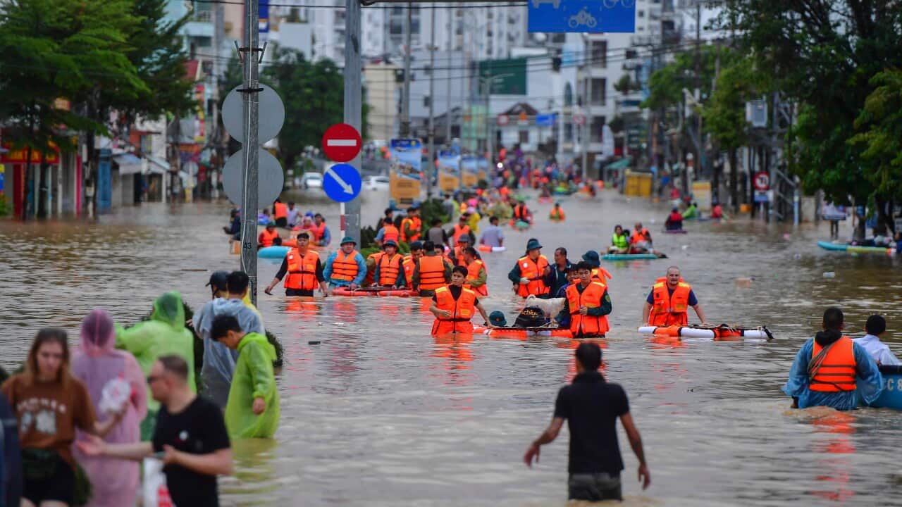 Dozens of people, some wearing high vis life jackets, wading through waist heigh brown flood waters on a city street