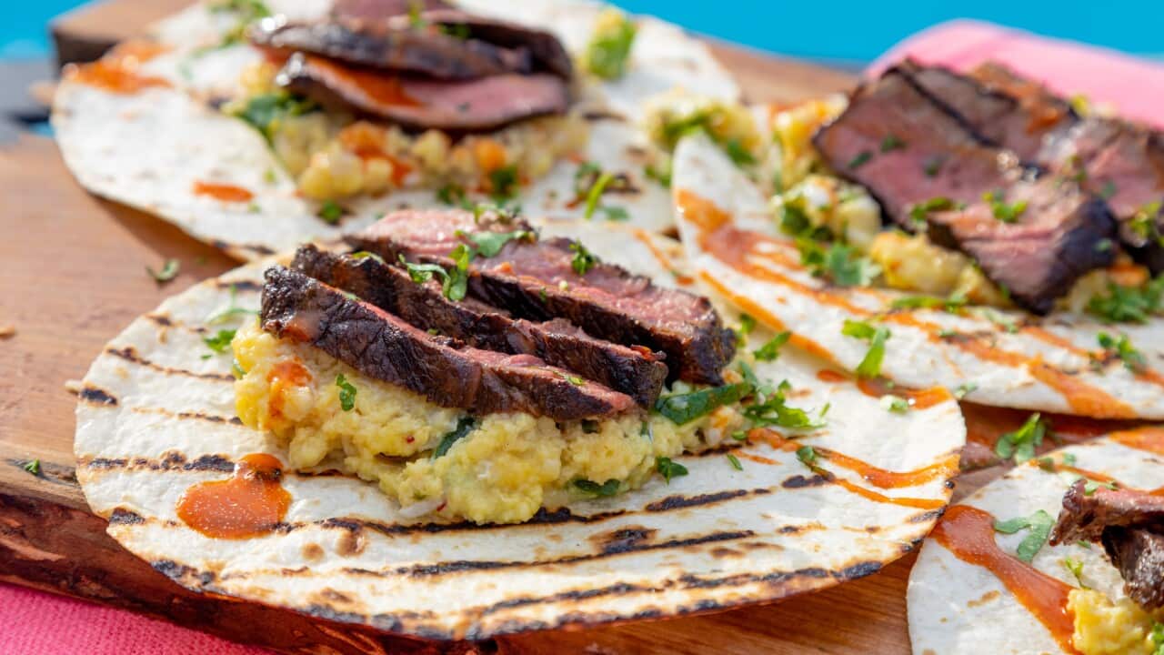 A row of flat tortillas with charry grill marks sit on a board, each with scrambled eggs, strips of steak, herbs and a drizzle of sauce.