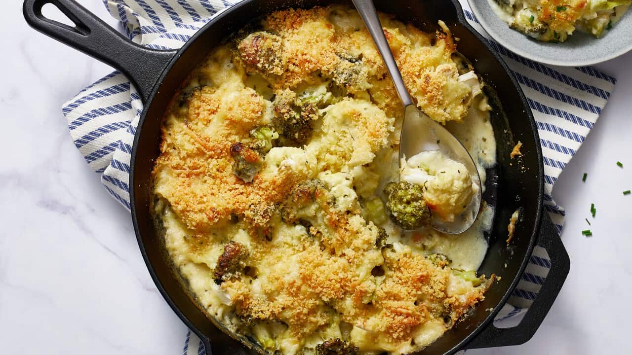 A cast iron skillet holds a creamy veg bake, topped with golden breadcrumbs. A spoon sits in the pan, and a serve of the vegetable bake sits in a bowl beside the skillet.