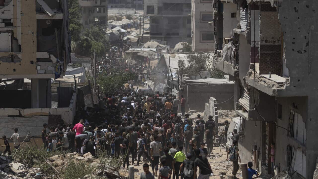 A large group of Palestinians walking down a pathway with heavily damaged buildings on either side.