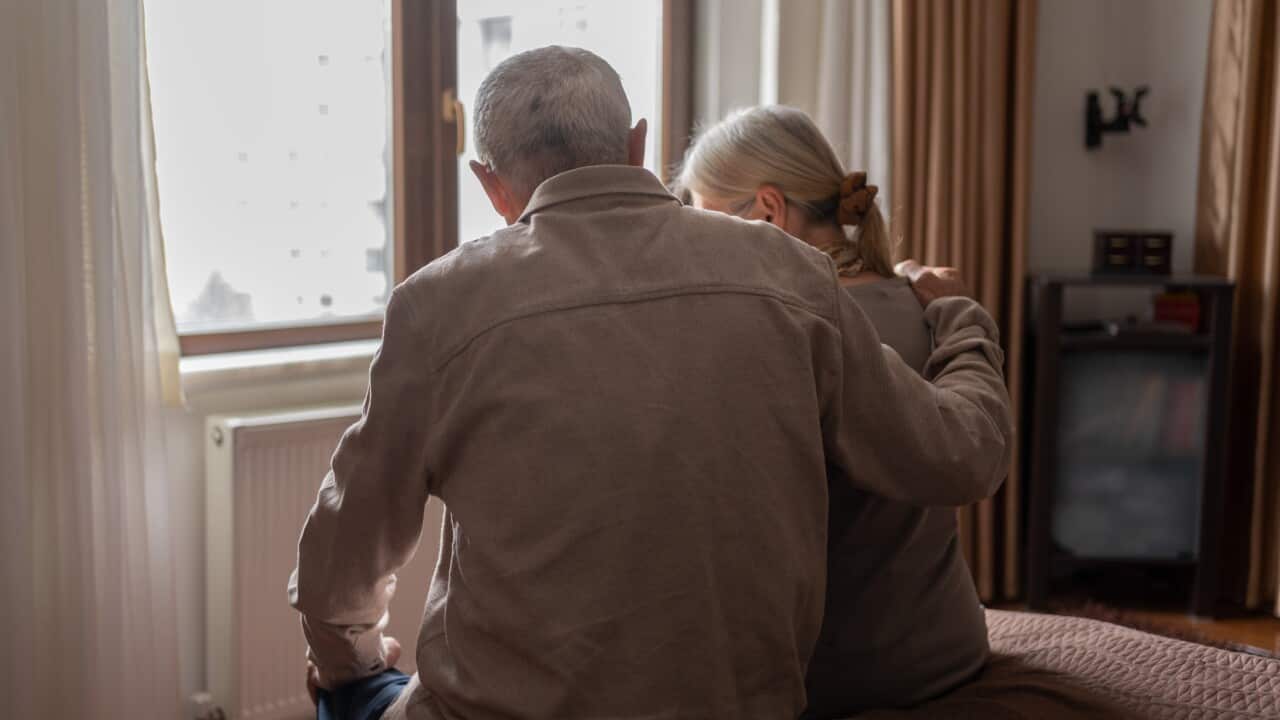 An older man and older woman sitting on a bed, with their backs turned. The older man is comforting the woman, putting his arm around her.