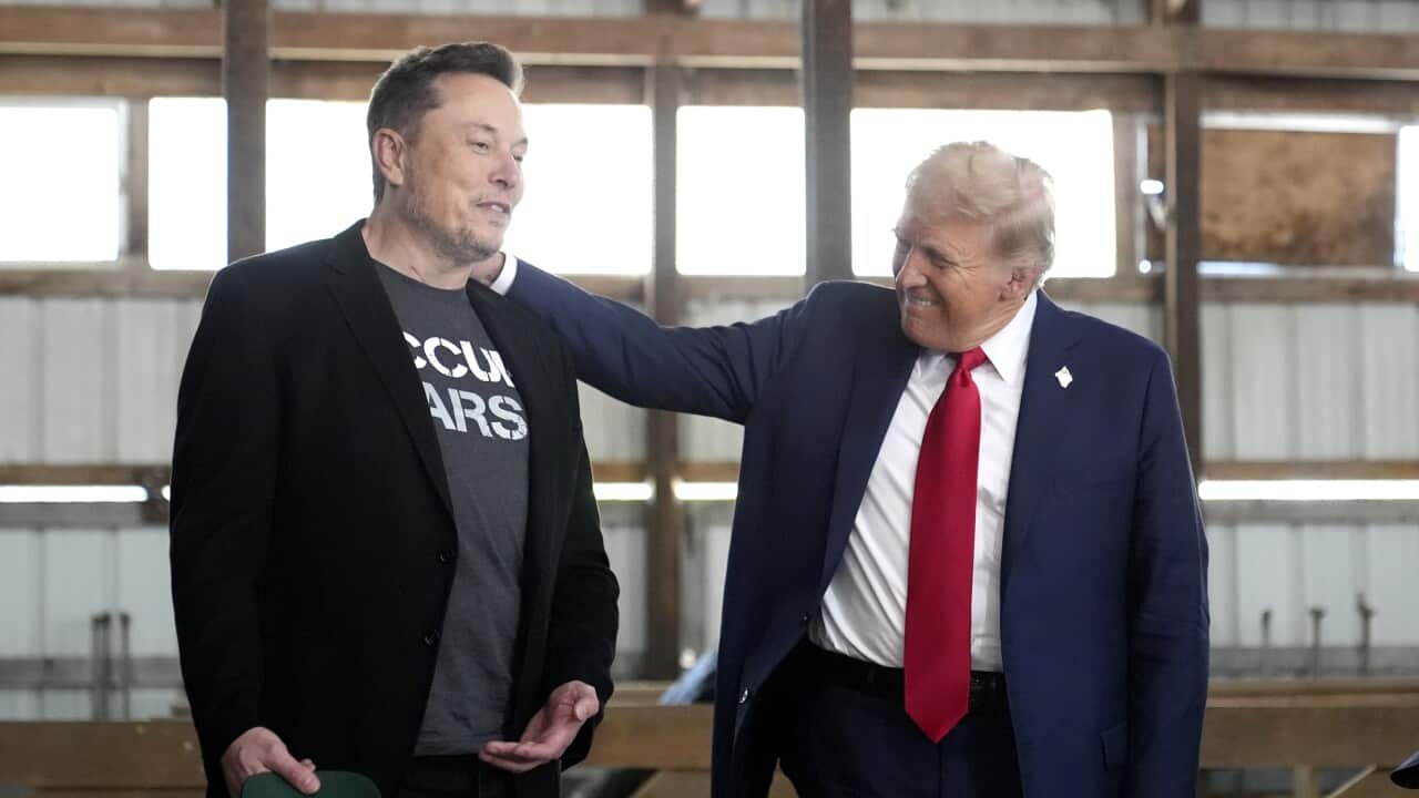 Donald Trump, wearing a dark blue suit and red tie, stands with his hand on the shoulder of Elon Musk, who is dressed in black.