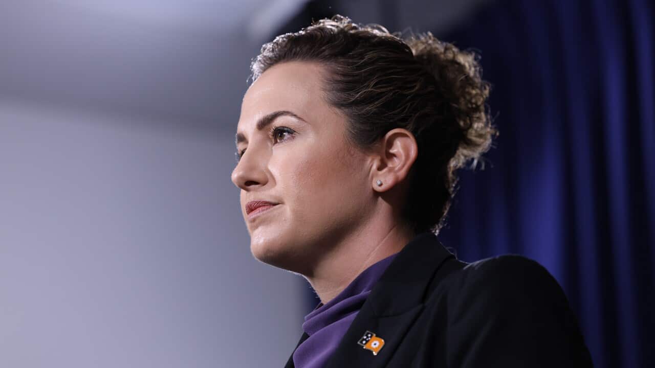 A woman wearing a black blazer, with her hair in a bun, standing in front of a blue curtain.