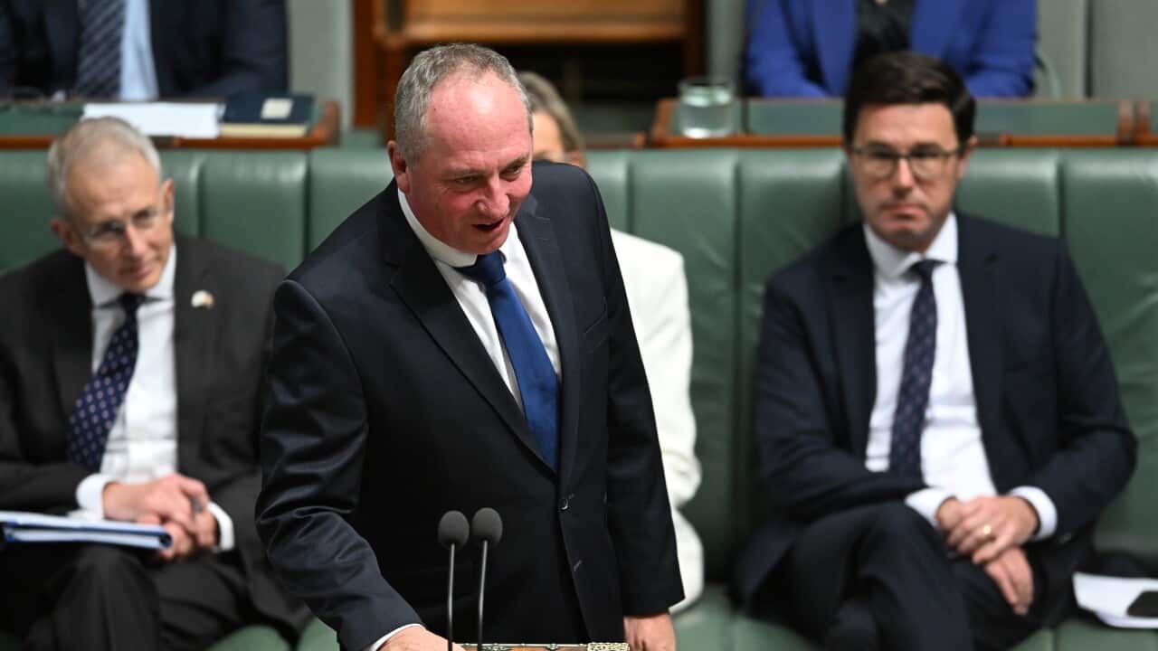 A man in a black suit stands behind a lectern, with two other men in black suits sitting behind him.
