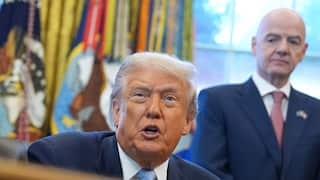 Donald Trump sitting in the Oval Office. A man is standing behind him in a blue blazer, white shirt and red tie.
