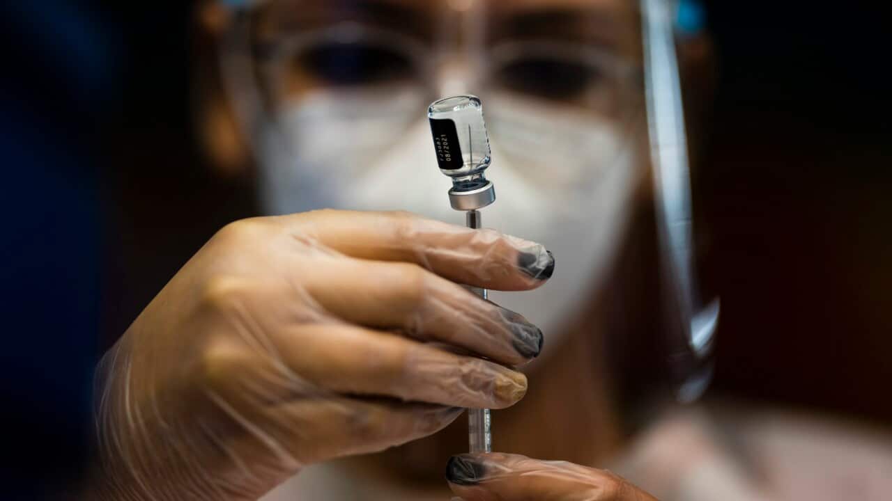 A scientist wearing a face mask, gloves and other personal protective equipment. They are holding a small glass bottle with a needle poking into the top.