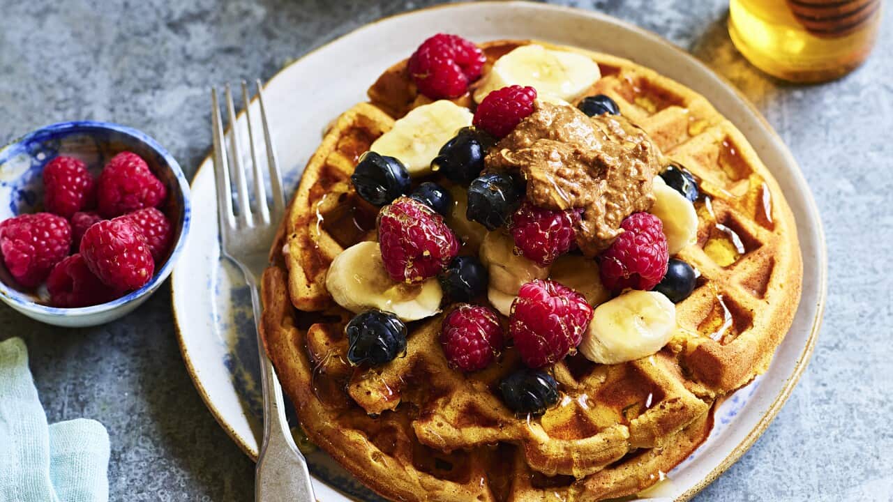 Two round waffles sit on a while plate on an speckled blue-grey surface. Berries, banana slices and syrup top the waffles. A cup of tea sits alongside the plate.