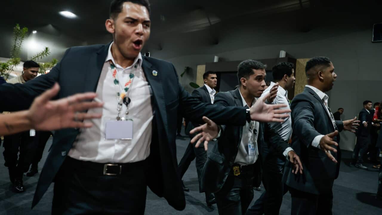 A row of men in suits and lanyards hold their arms out as they move through a large, dark indoor space.