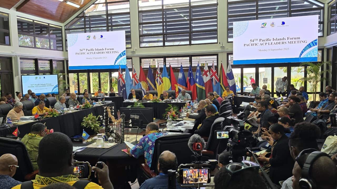 A wide-angle photo shows a large indoor meeting with a group of people seated at long tables.