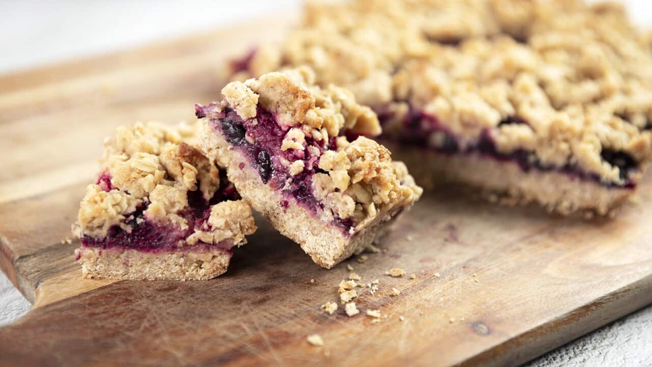 Two pieces of a three layer slice, with a thick biscuit bottom, jammy-looking filling and crumble topping, sit in front of a larger slab of the slice, all on a wooden board.