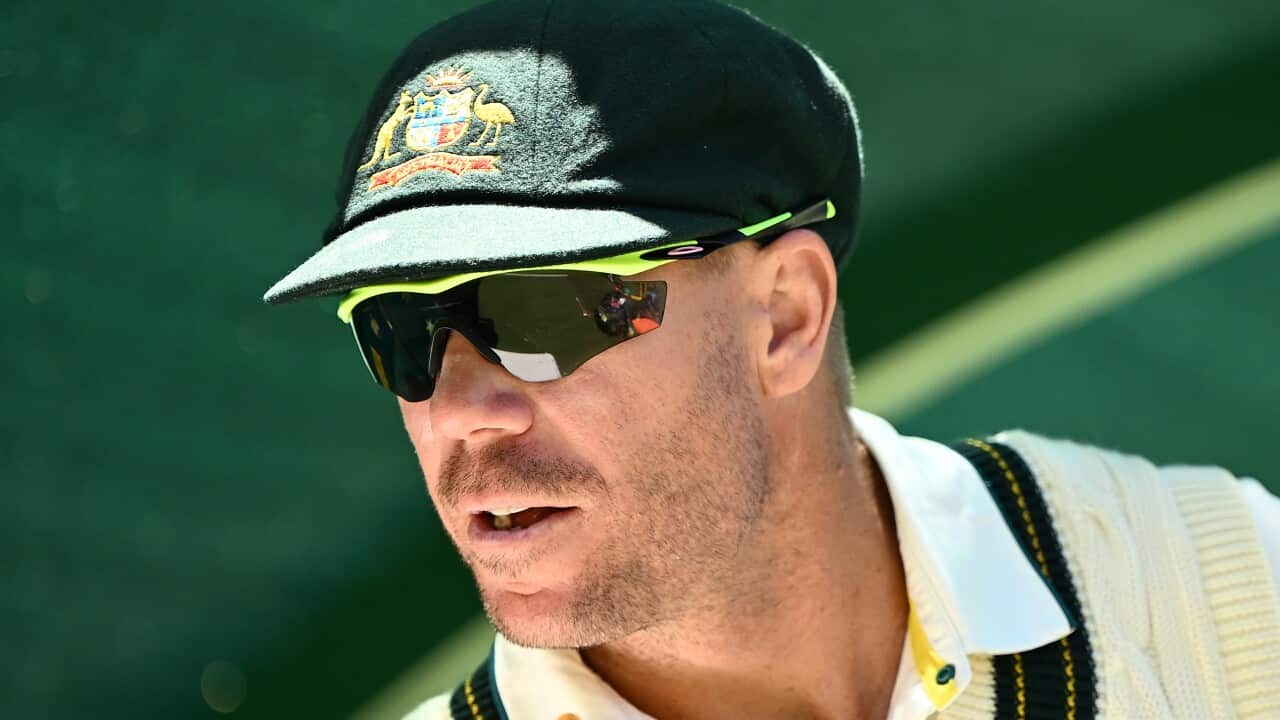 A close-up of a man wearing a green cap, a white cricketer's vest and sunglasses.