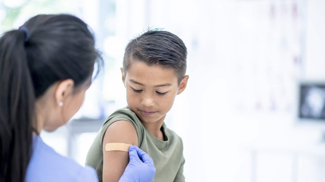 A doctor wearing blue scrubs and blue gloves places a band-aid on a young boy's arm.